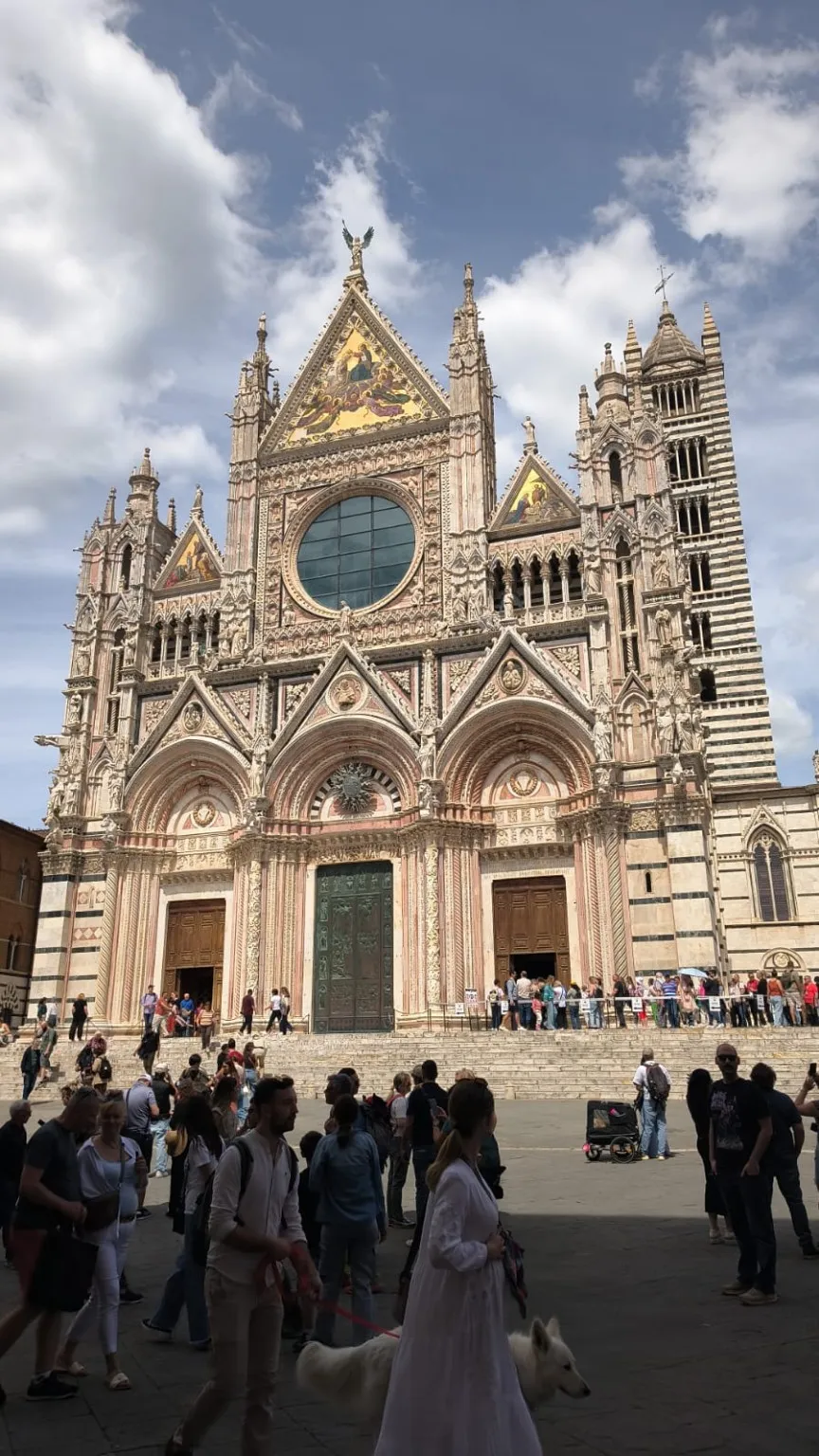Façade du Duomo de Sienne vue depuis la Piazza del Duomo, avec des touristes sur les marches et un ciel nuageux.